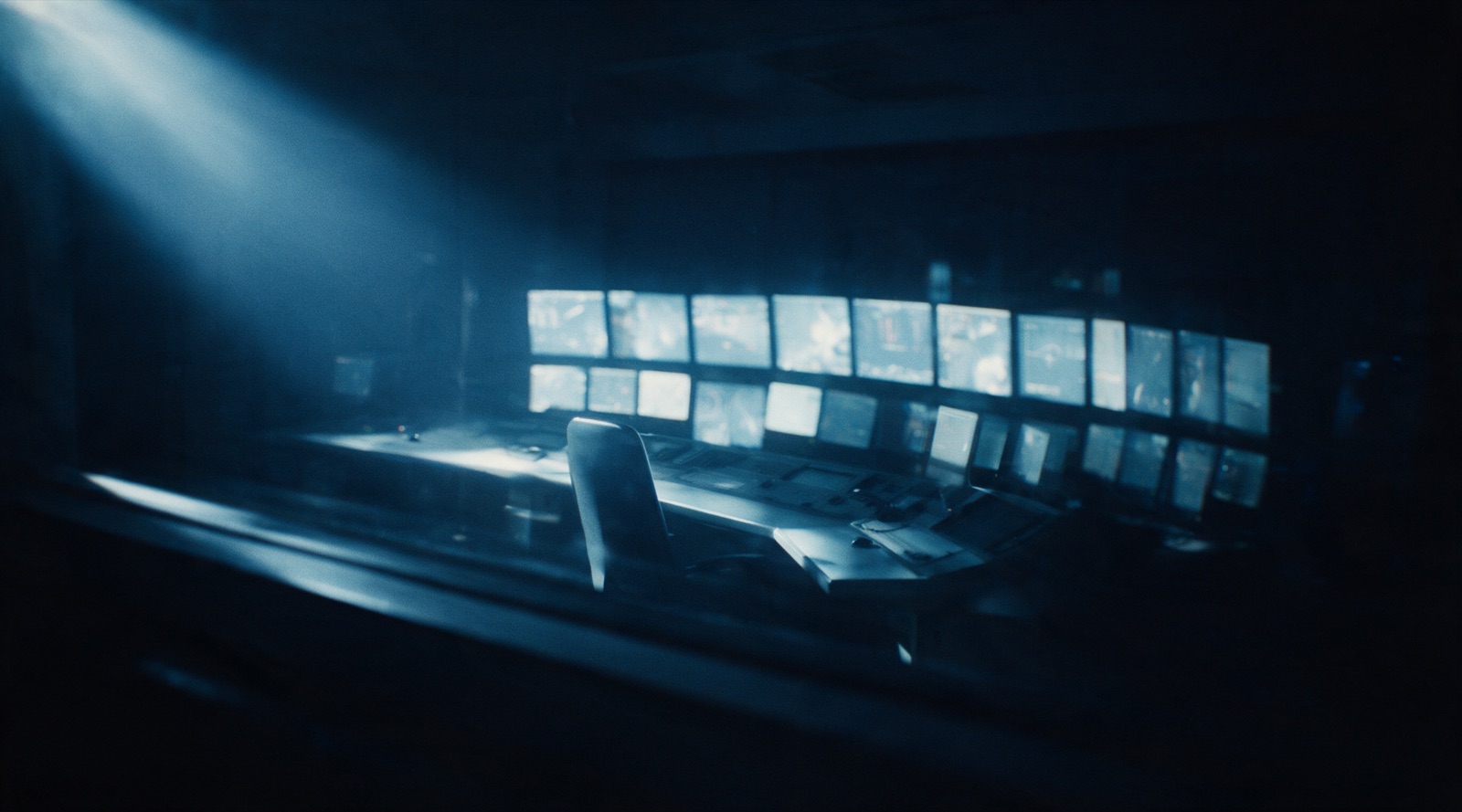 Sala de controle vazia à noite: um painel de monitores como única fonte de luz e uma cadeira desocupada diante do console.